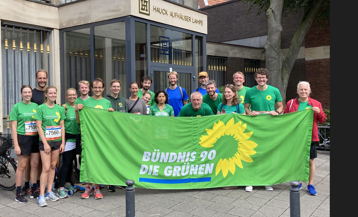 Eine Gruppe von Menschen hat vor sich ein Banner aufgestellt. Das Banner hat einen grünen Hintergrund, auf dem das Logo der Partei BÜNDNIS 90/DIE GRÜNEN zu sehen ist: Der Name Die GRÜNEN in weißer Schrift, dazu eine gelbe Sonnenblume.